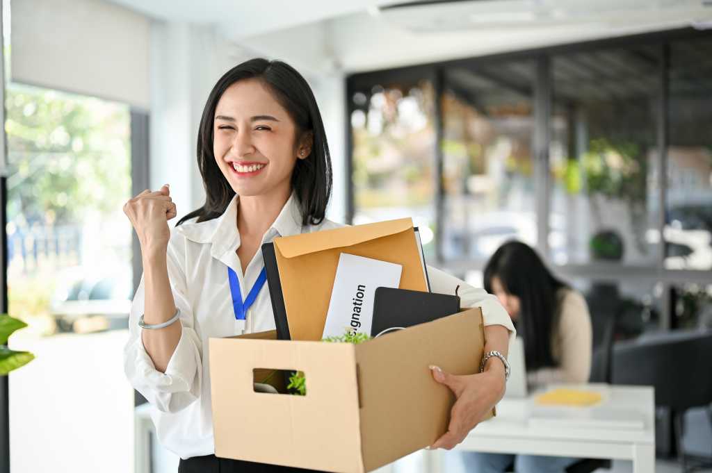 Smiling and excited young Asian female office worker is celebrating her resignation, carrying her personal stuff, happy to leave her job or change job positions.
